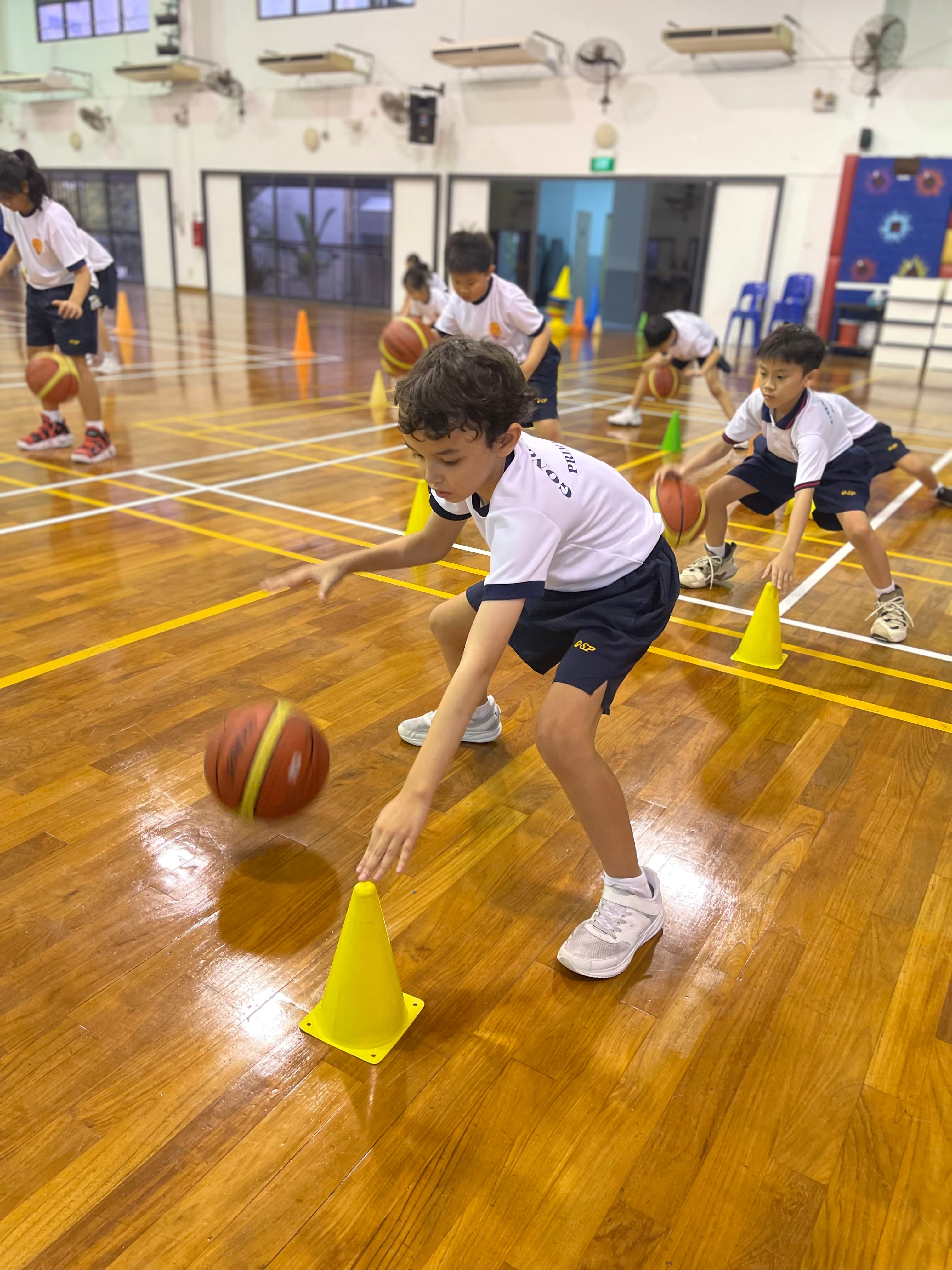 Children in school uniforms dribble basketballs around yellow cones on a wooden indoor court.